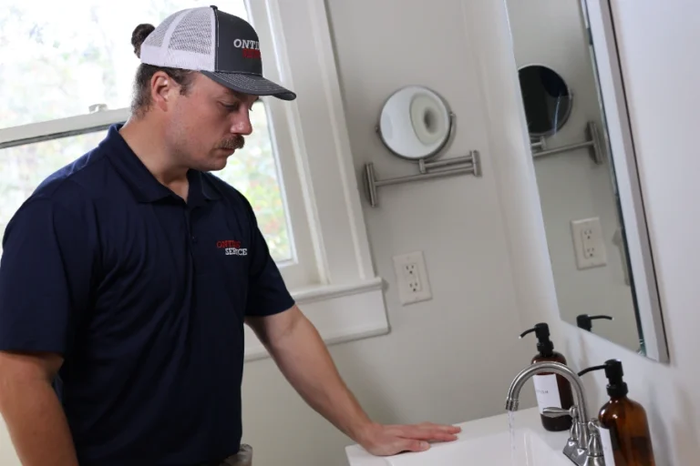 A man in a navy shirt and baseball cap stands by a bathroom sink with running water, looking down at the counter.