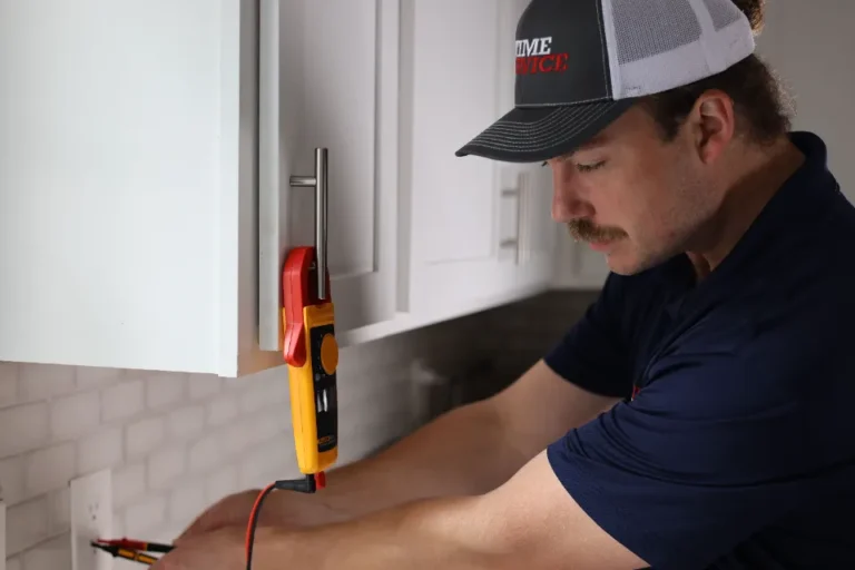 A man in a cap uses a digital multimeter to test an electrical outlet in a kitchen with white cabinets and a tiled backsplash.