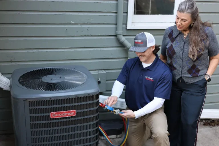 A technician inspects and adjusts gauges on a Goodman air conditioning unit outside a house while a woman stands nearby watching.