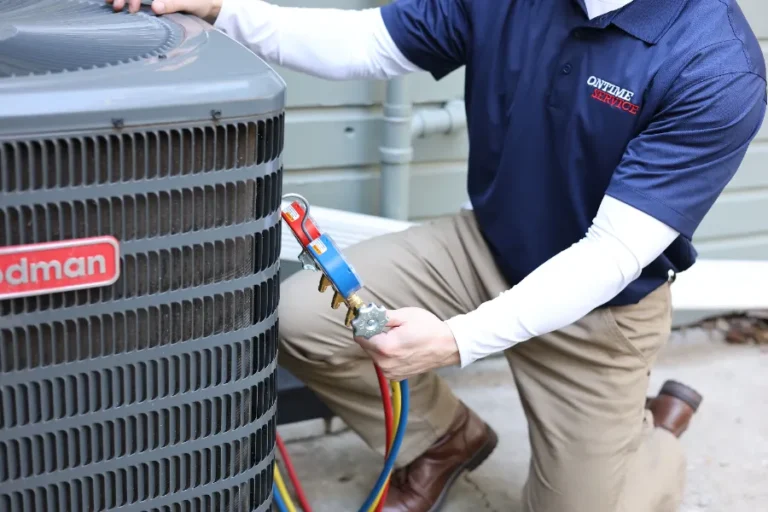 A technician in khaki pants and a blue shirt services an outdoor Goodman air conditioning unit using a diagnostic gauge with colored hoses.