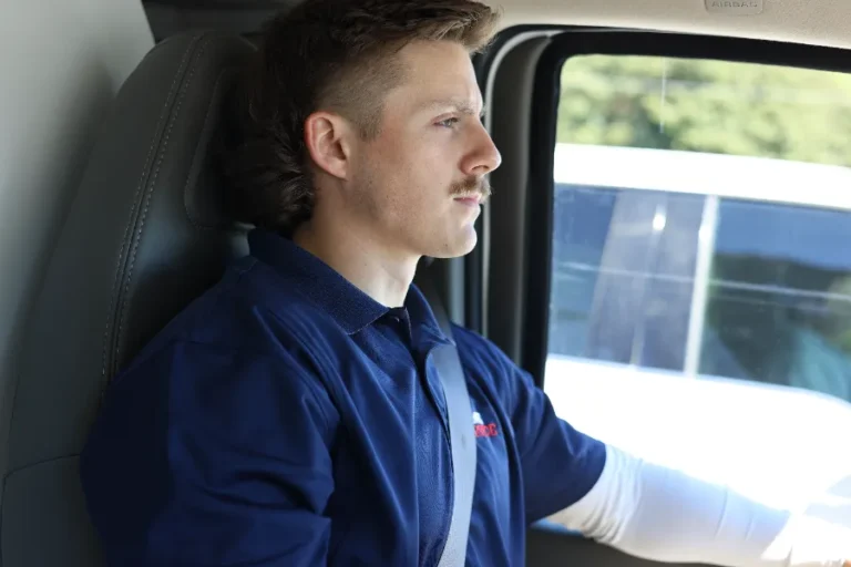 A man in a blue collared shirt and seatbelt is driving a vehicle, looking straight ahead. Sunlight comes through the window.