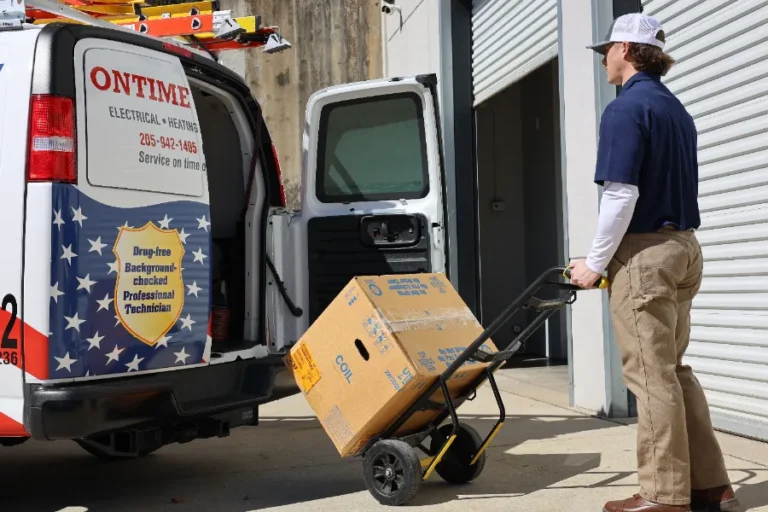 A worker in a blue shirt and cap uses a hand truck to load a cardboard box into an OnTime Electrical van parked near a building with open garage doors.