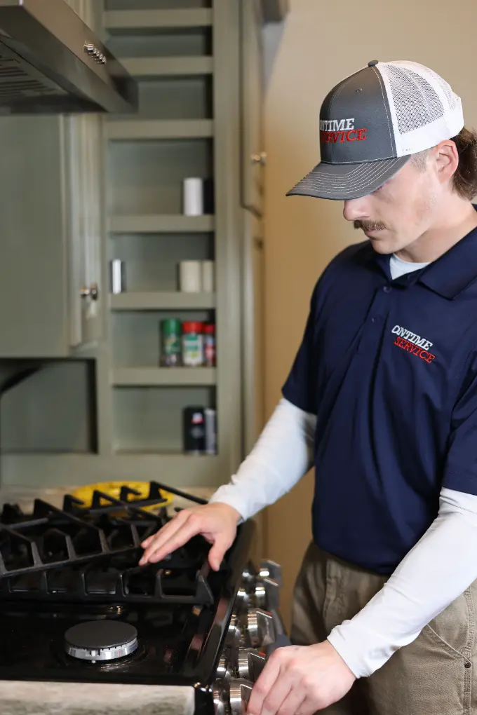 A technician in a uniform and cap stands in a kitchen, inspecting or adjusting a gas stove with his hands on the stove's controls.