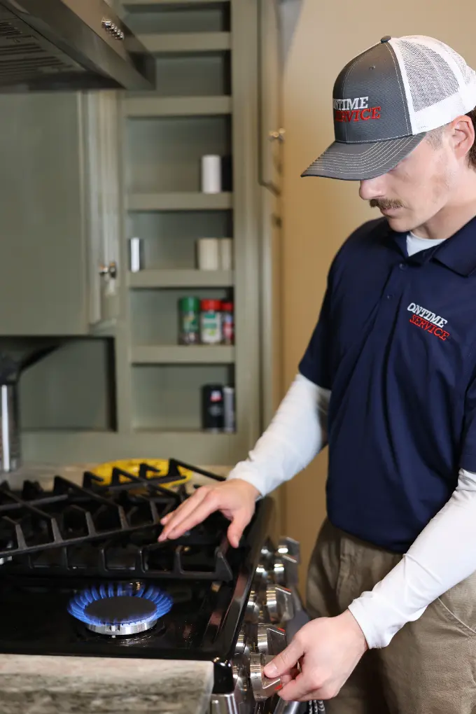 A technician in uniform adjusts the dial on a gas stove with a blue flame on, standing in a kitchen near open shelves with spice containers.