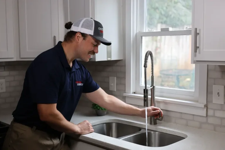 A man wearing a uniform and cap tests the water flow from a kitchen sink faucet in a modern, well-lit kitchen.