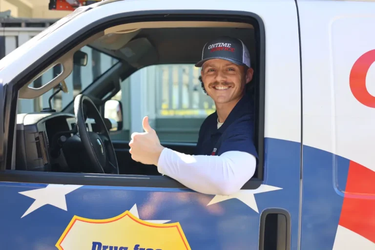 A man wearing a cap and navy shirt sits in a vehicle’s driver seat, smiling and giving a thumbs up.