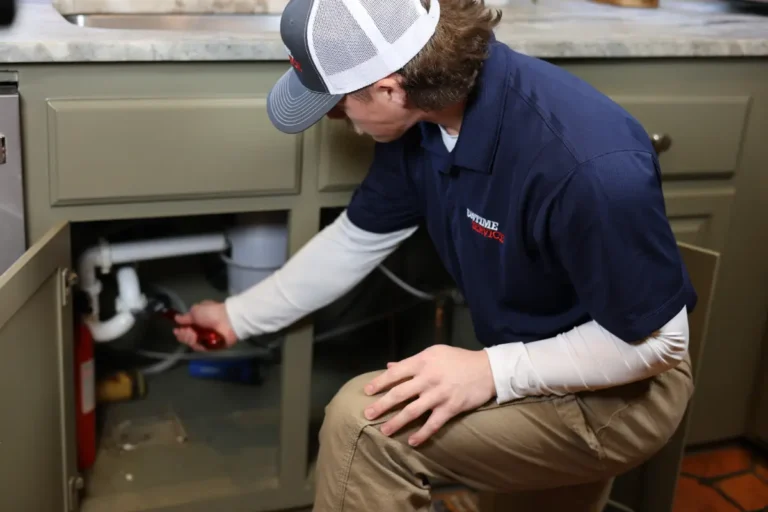 A plumber in a navy shirt and cap inspects pipes under a kitchen sink, holding a flashlight and kneeling in front of an open cabinet.