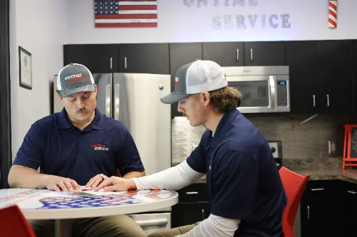Two men in matching uniforms and hats sit at a round table in a break room with an American flag on the wall behind them.