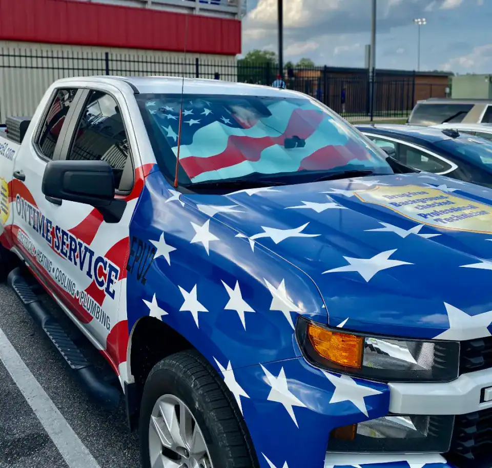 Pickup truck with an American flag-themed wrap advertises an HVAC and plumbing service; parked in a lot on a cloudy day.
